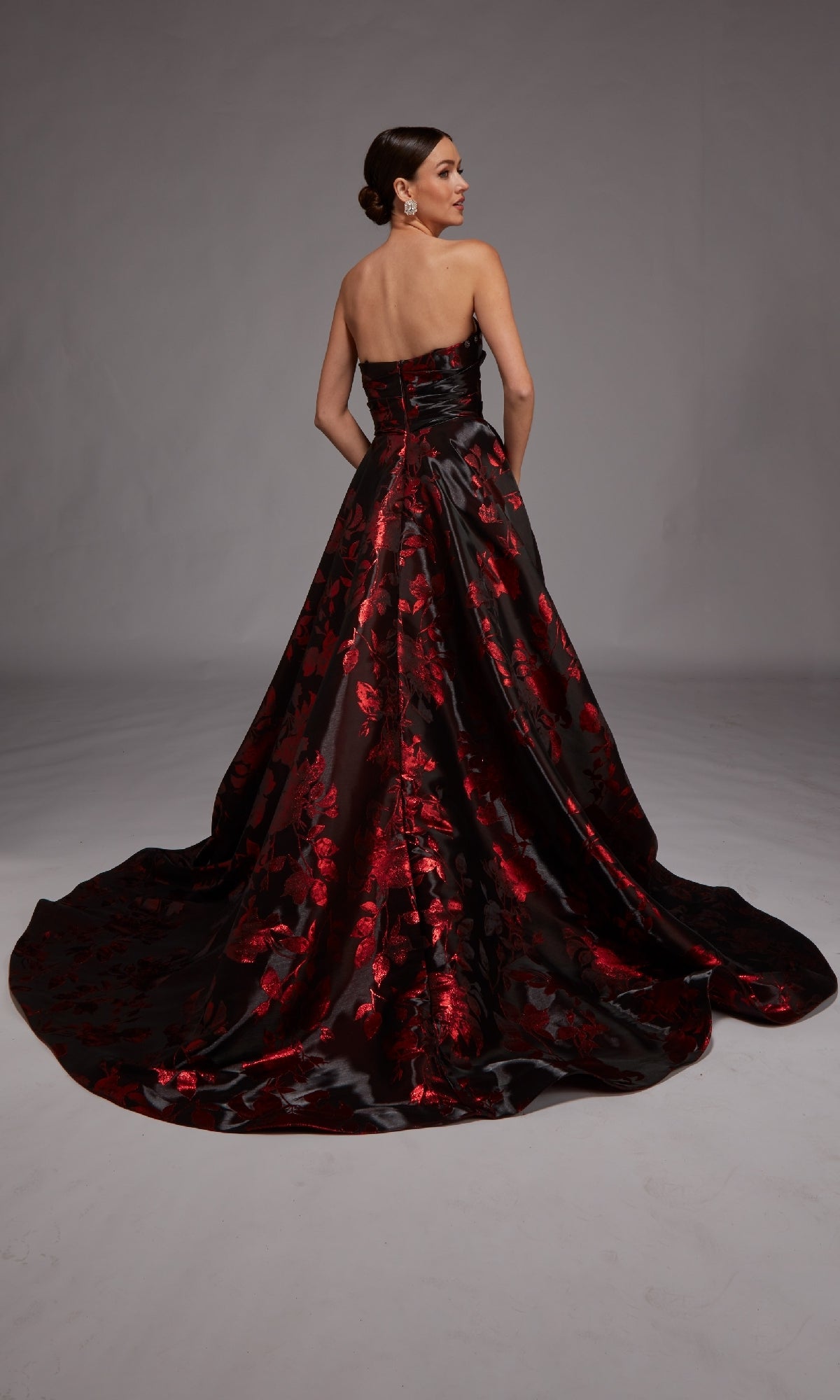 A woman faces away from the camera in the Alyce 27862, a strapless black floral jacquard gown with a red pattern, floor-length silhouette, and long train.
